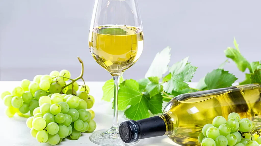 Ly rượu vang trắng vàng nhạt bên chai vang nằm nghiêng và những chùm nho xanh trên nền sáng