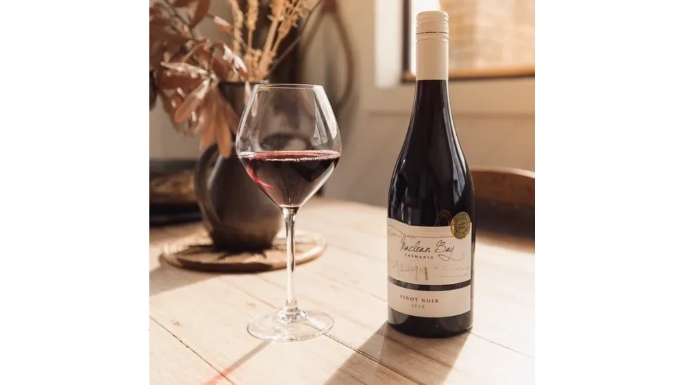 Chai vang Maclean Bay Pinot Noir cùng ly vang đỏ đặt trên bàn gỗ sáng cạnh bình hoa khô